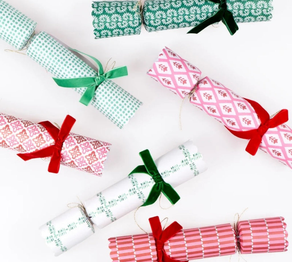 Six Christmas cracker ornaments wrapped in festive patterned paper and tied with green and red ribbons, arranged on a clean white background.