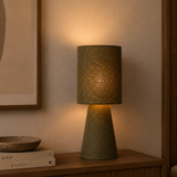 A textured table lamp with a woven drum shade and tapered base in a warm olive-green tone, glowing with a soft yellow light on a wooden nightstand in a cozy bedroom setting.
