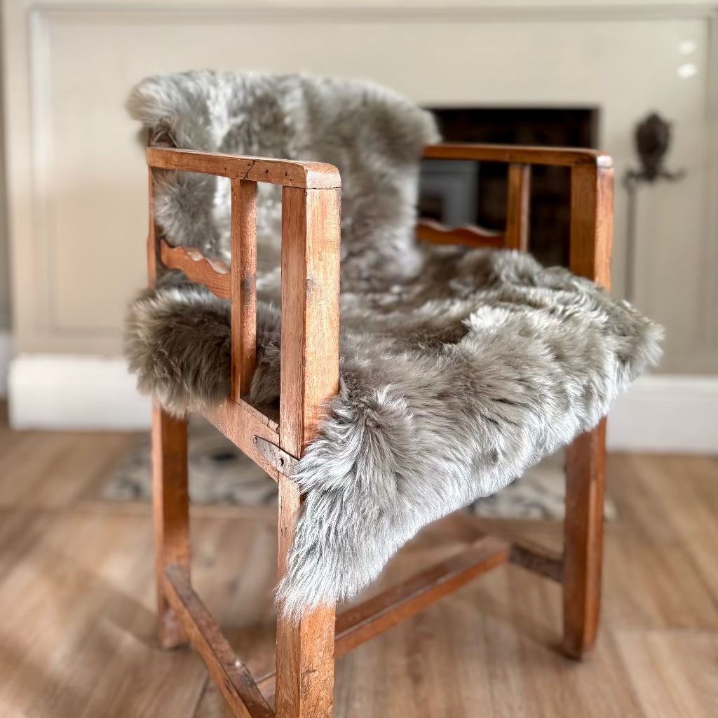 A rustic wooden armchair with a thick, fluffy gray sheepskin or faux-fur seat and back cushion, set in a warm, neutral-toned room with a fireplace in the background.