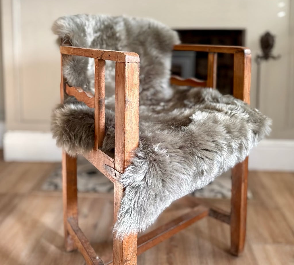 A rustic wooden armchair with a thick, fluffy gray sheepskin or faux-fur seat and back cushion, set in a warm, neutral-toned room with a fireplace in the background.