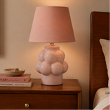 A pink ceramic table lamp with a textured bubble base and a matching pink fabric drum shade, styled on a wooden nightstand in a bedroom setting.