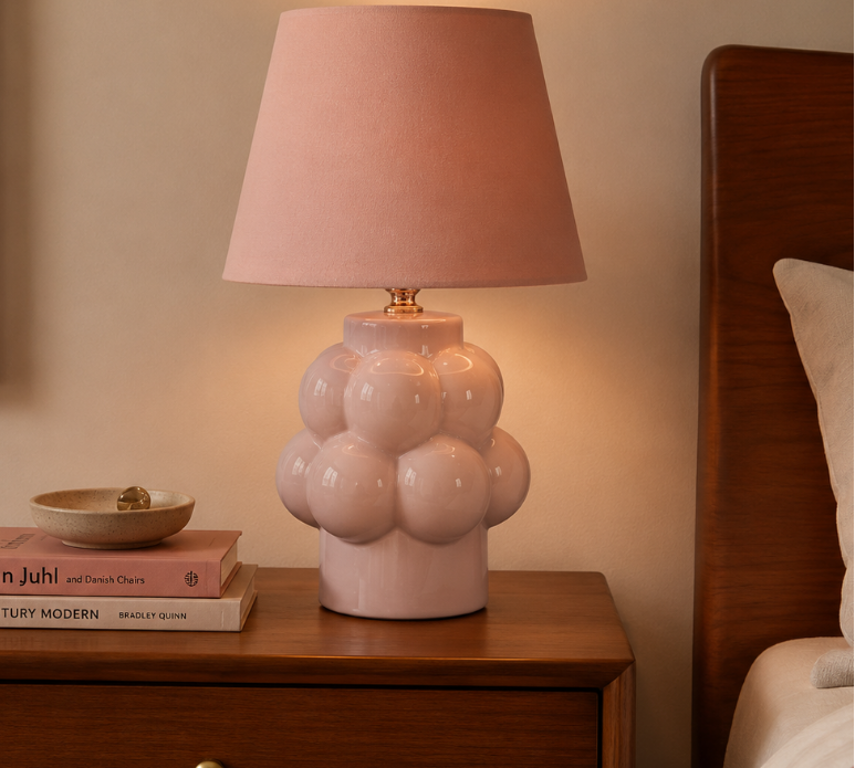 A pink ceramic table lamp with a textured bubble base and a matching pink fabric drum shade, styled on a wooden nightstand in a bedroom setting.