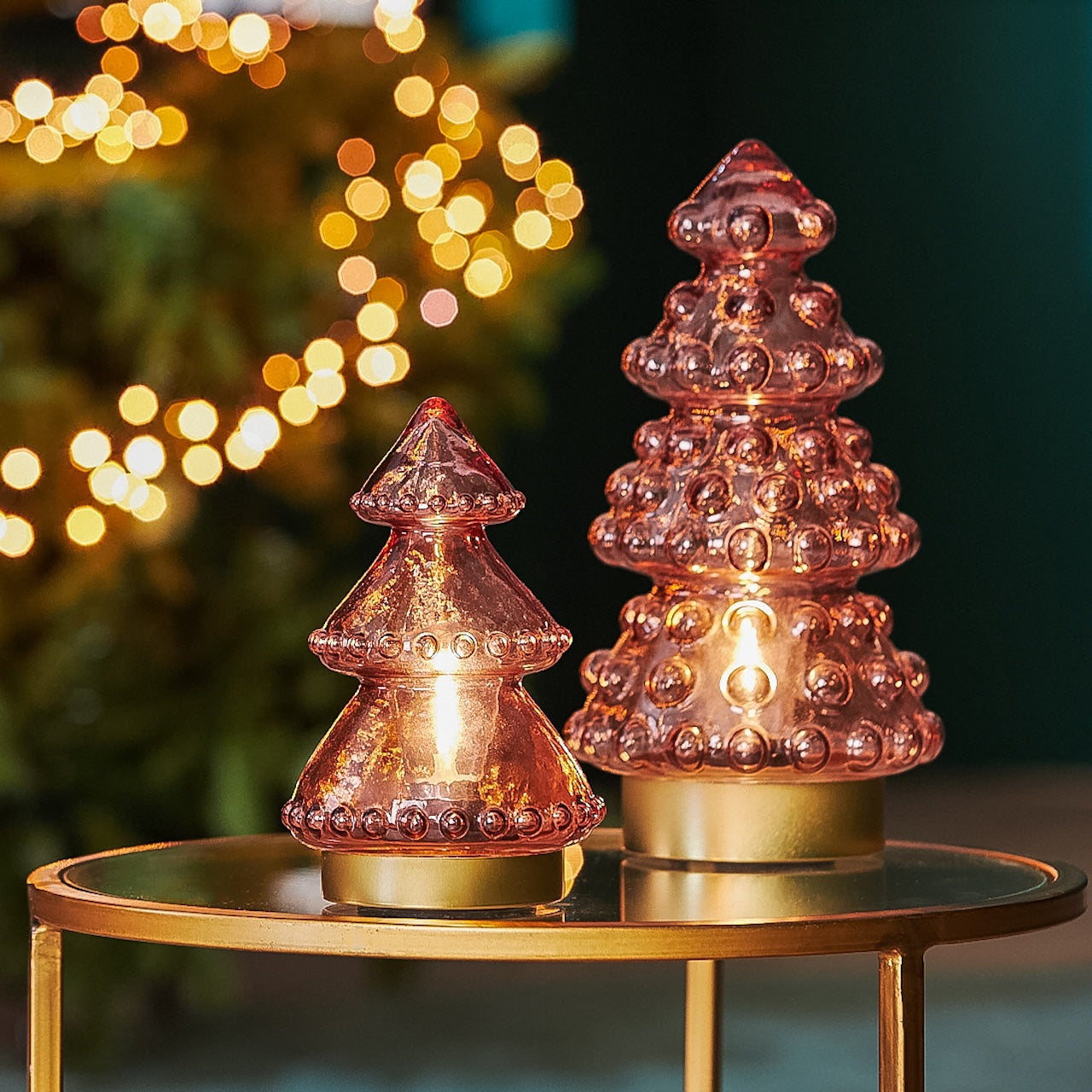 Two pink glass Christmas tree lights on a gold table with blurred lights in the background