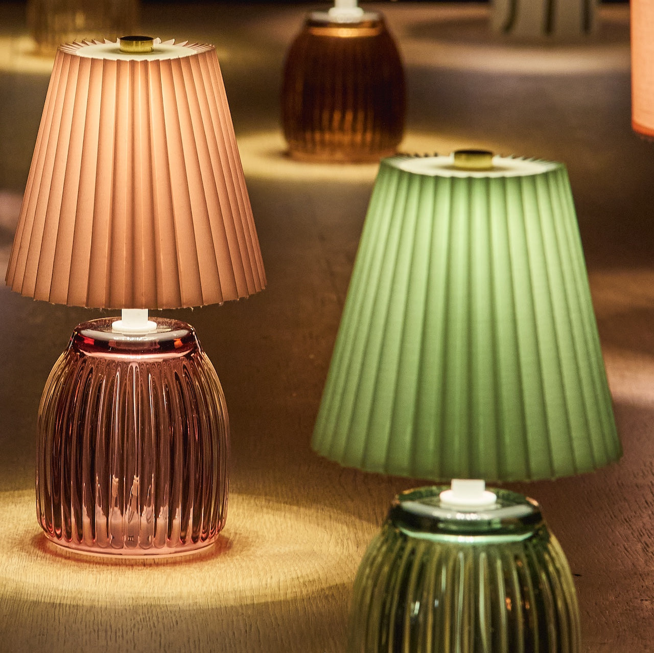 Two table lamps with pleated shades, one pink and one green, on a textured surface.