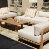Outdoor patio furniture set featuring a light wood-framed sectional sofa with off-white cushions, a matching armchair, a wooden coffee table, a closed cream patio umbrella, and a beige building in the background.