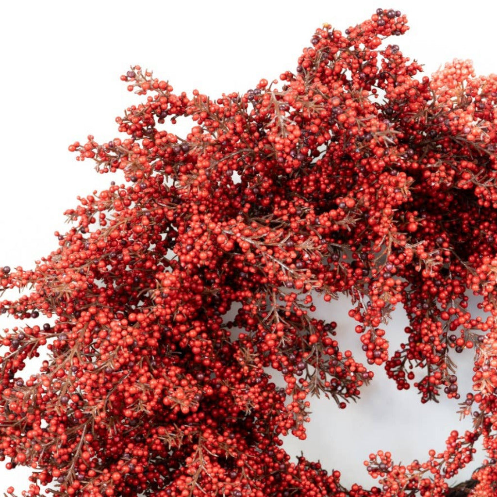 A close-up of a decorative wreath or floral arrangement covered in dense clusters of small red berries with muted brown foliage, shown against a plain white background.