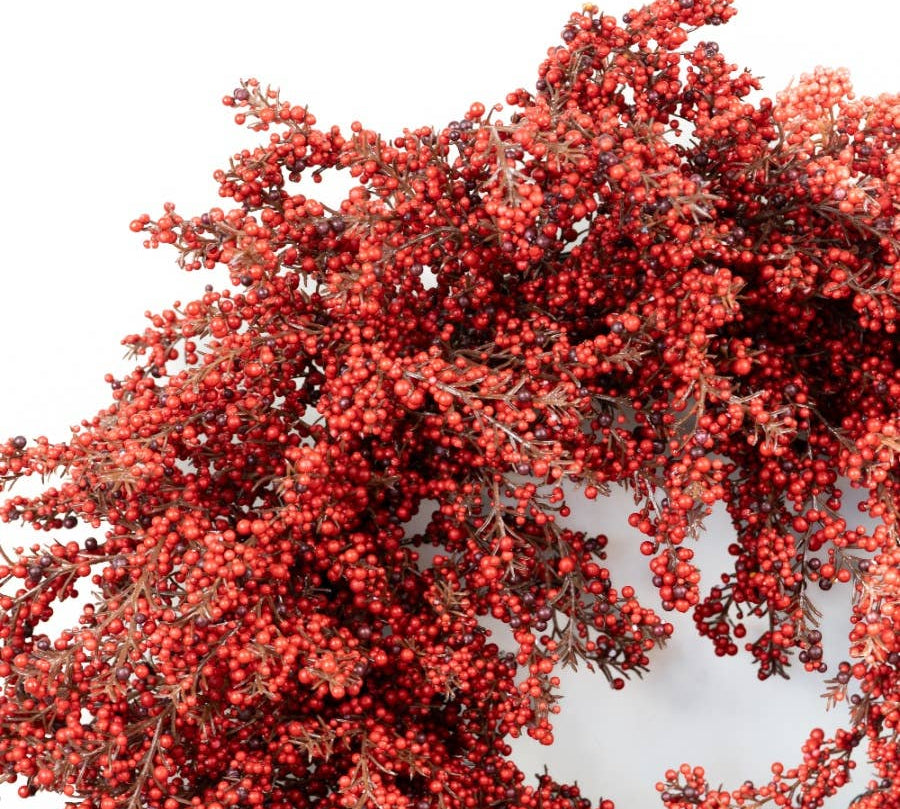 A close-up of a decorative wreath or floral arrangement covered in dense clusters of small red berries with muted brown foliage, shown against a plain white background.