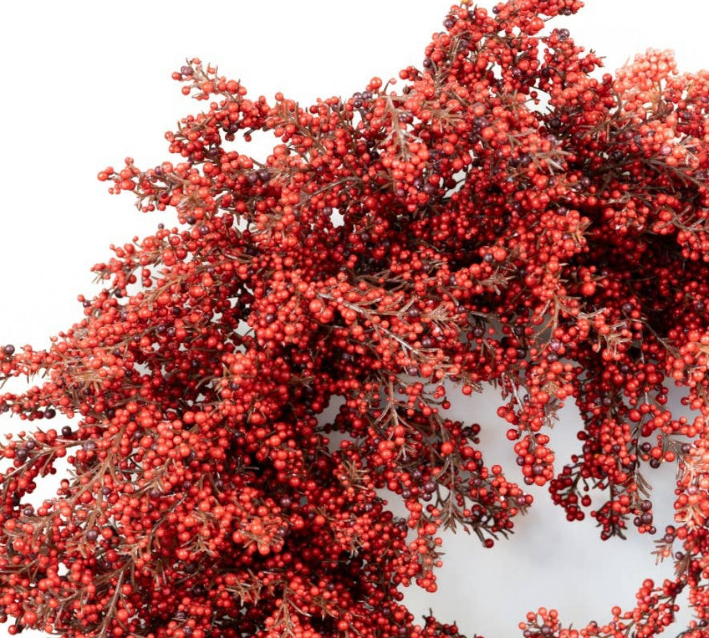 A close-up of a decorative wreath or floral arrangement covered in dense clusters of small red berries with muted brown foliage, shown against a plain white background.