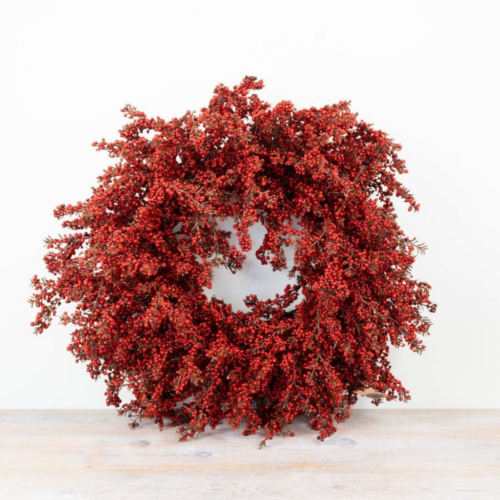 A round artificial berry wreath in vibrant red, displayed on a light wood surface against a plain white background.