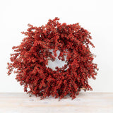 A round artificial berry wreath in vibrant red, displayed on a light wood surface against a plain white background.