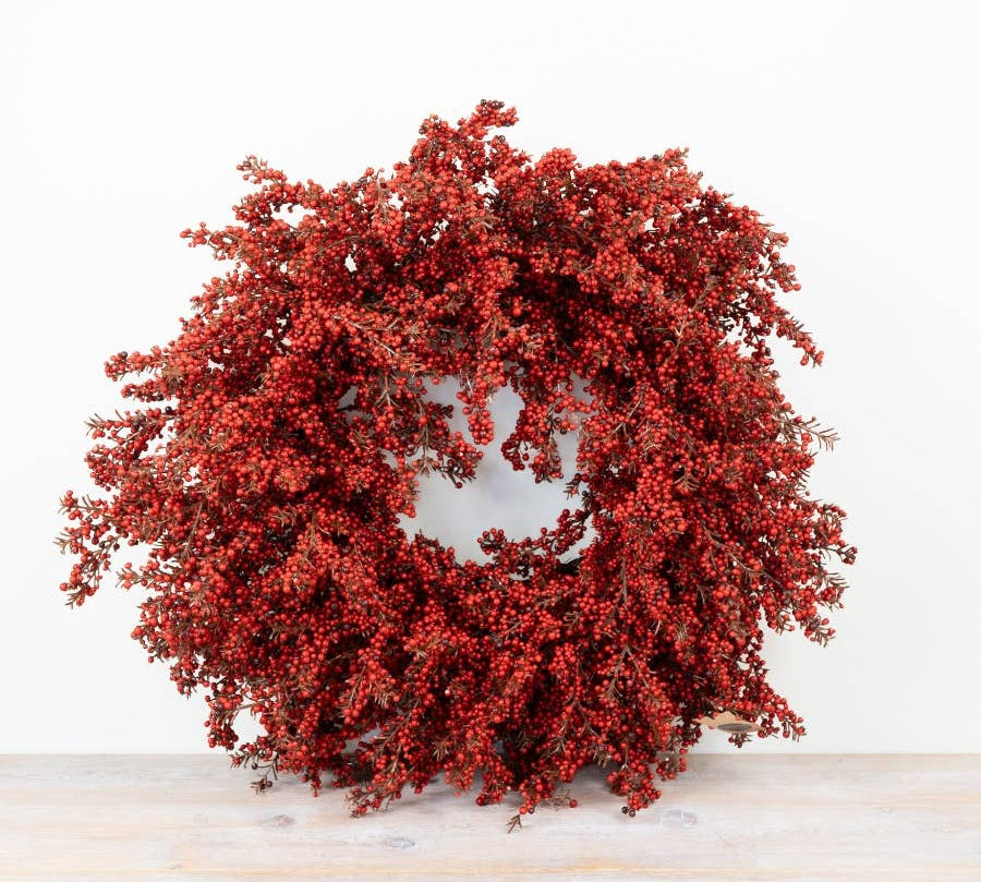 A round artificial berry wreath in vibrant red, displayed on a light wood surface against a plain white background.