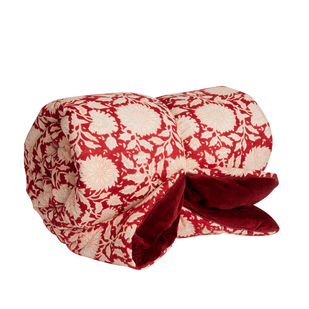 A rolled-up quilted blanket featuring a red and cream floral pattern on the exterior and a plush dark red velvet lining visible on the underside, set against a plain white background.
