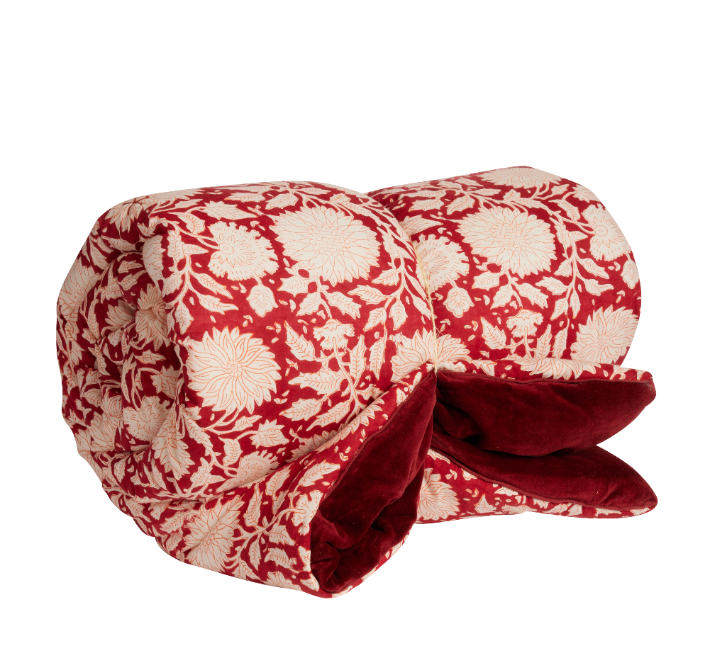 A rolled-up quilted blanket featuring a red and cream floral pattern on the exterior and a plush dark red velvet lining visible on the underside, set against a plain white background.