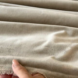 A close-up of a velvet fabric with a soft, brushed texture, draped in horizontal folds with a hand holding the bottom edge to show the weave and fringe.