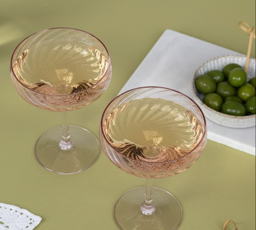 Two twisted champagne coupe glasses with a pale pink tint and textured spiral pattern, filled with white wine, set on a green tablecloth with a small bowl of olives in the background.