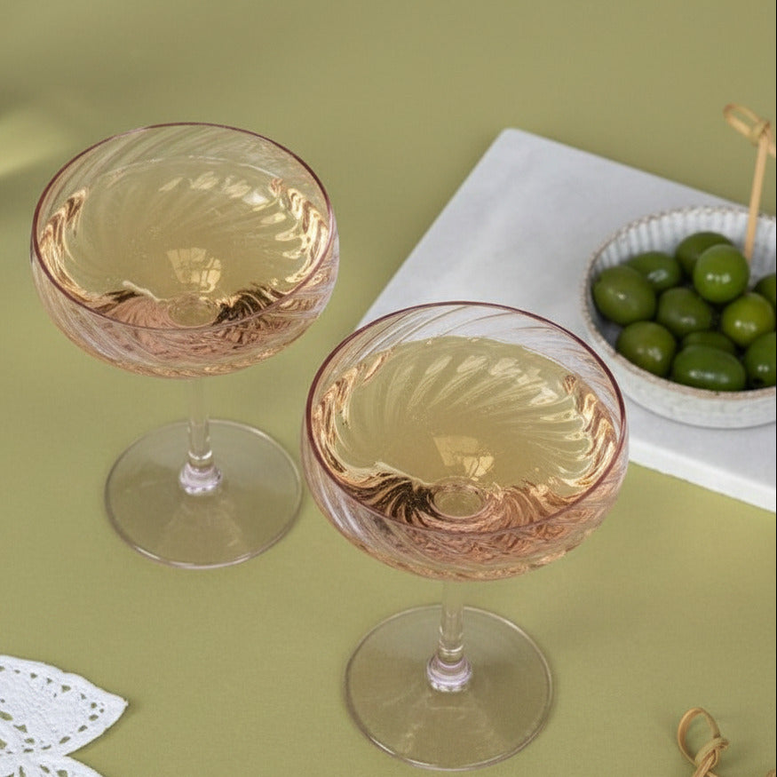 Two twisted champagne coupe glasses with a pale pink tint and textured spiral pattern, filled with white wine, set on a green tablecloth with a small bowl of olives in the background.