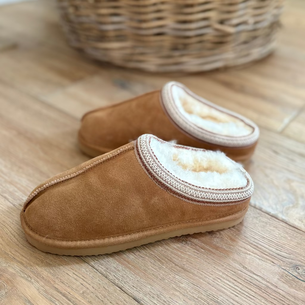 A pair of tan suede slip-on slippers with a soft cream shearling lining, displayed on a wooden floor with a woven basket in the background.