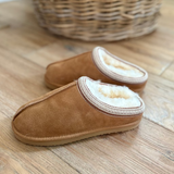 A pair of tan suede slip-on slippers with a soft cream shearling lining, displayed on a wooden floor with a woven basket in the background.