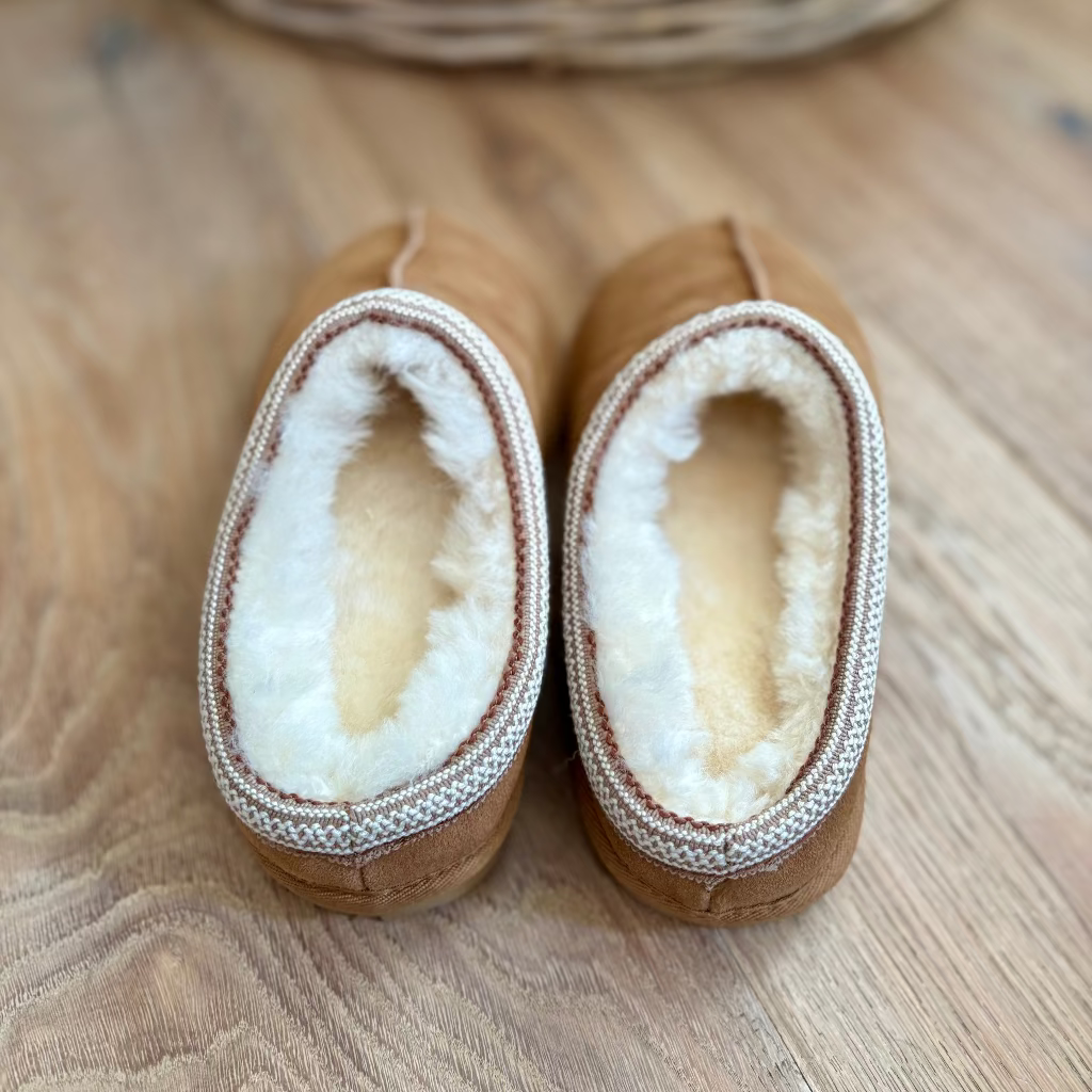 A pair of tan slip-on slippers with thick white shearling lining and a braided trim, shown from above on a wooden floor.