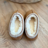 A pair of tan slip-on slippers with thick white shearling lining and a braided trim, shown from above on a wooden floor.