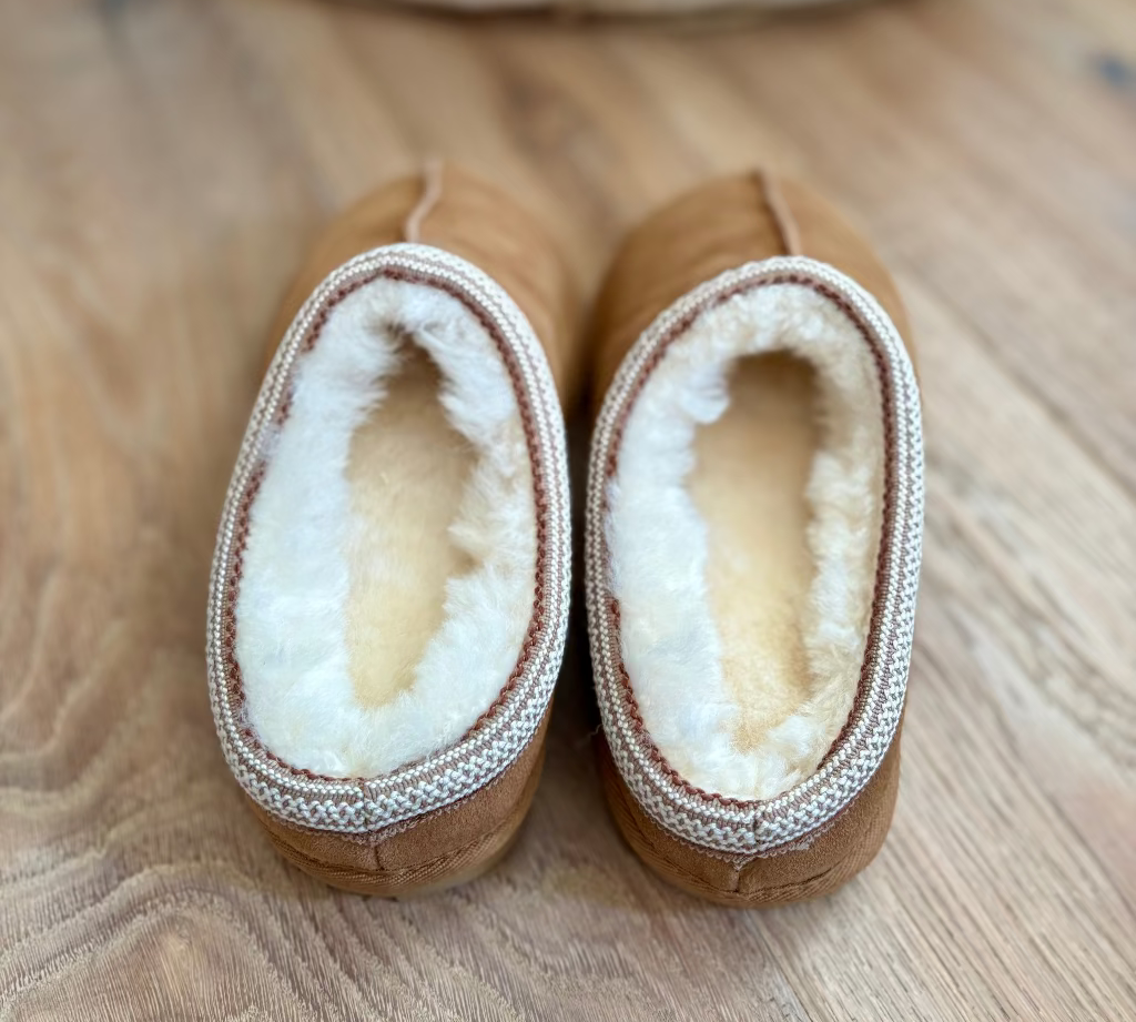 A pair of tan slip-on slippers with thick white shearling lining and a braided trim, shown from above on a wooden floor.