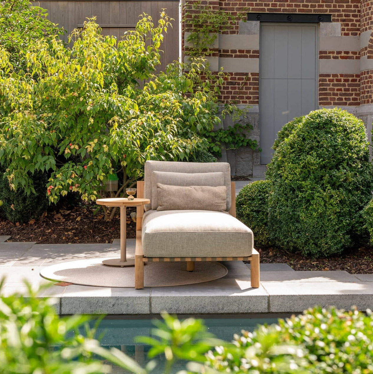 Grey lounger with wooden legs on a patio surrounded by greenery and a building.