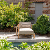 Grey lounger with wooden legs on a patio surrounded by greenery and a building.