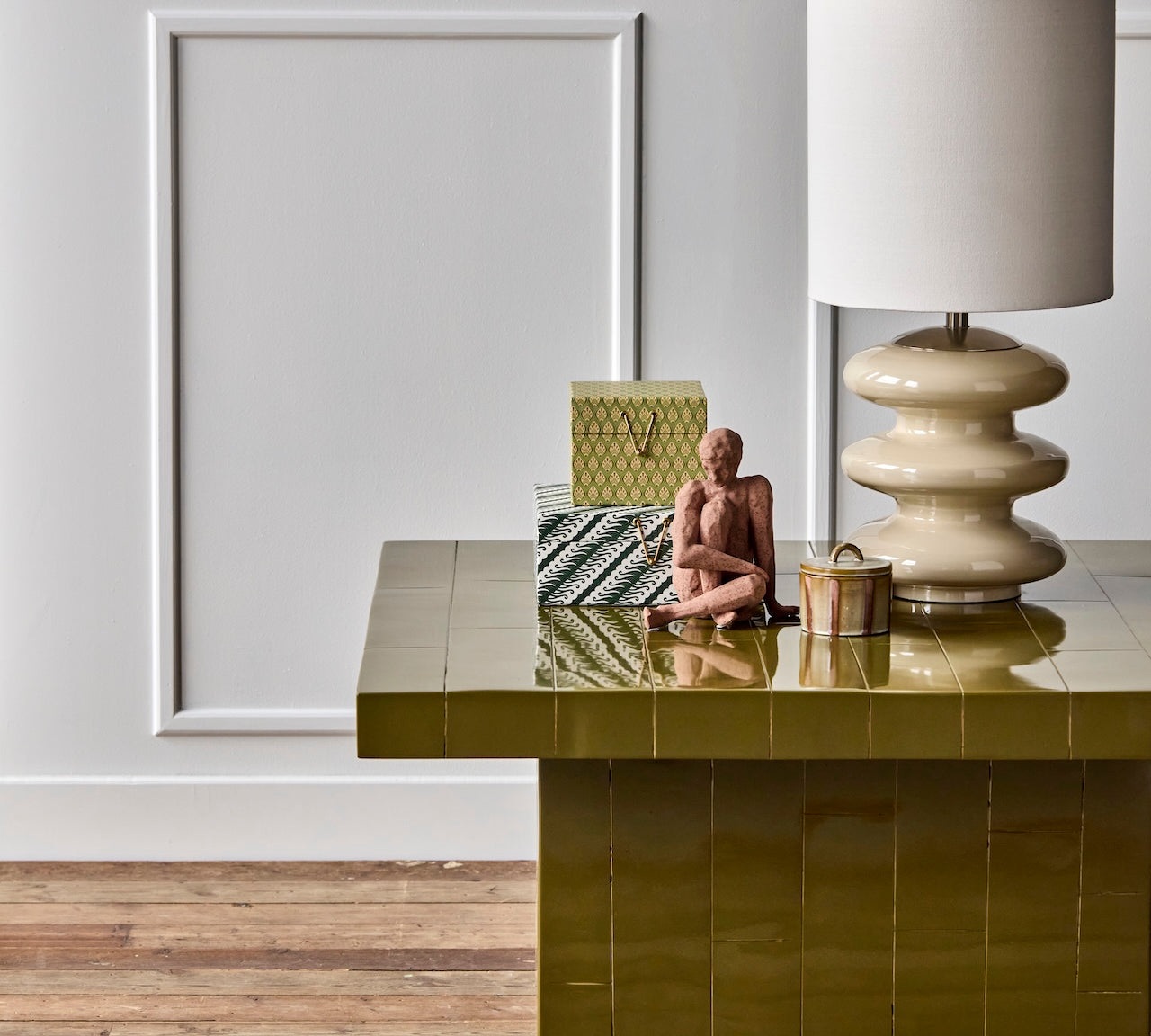 A glossy olive green tiled coffee table in a styled room setting, styled with a lamp, decorative boxes, and a small figurine.