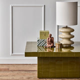 A glossy olive green tiled coffee table in a styled room setting, styled with a lamp, decorative boxes, and a small figurine.