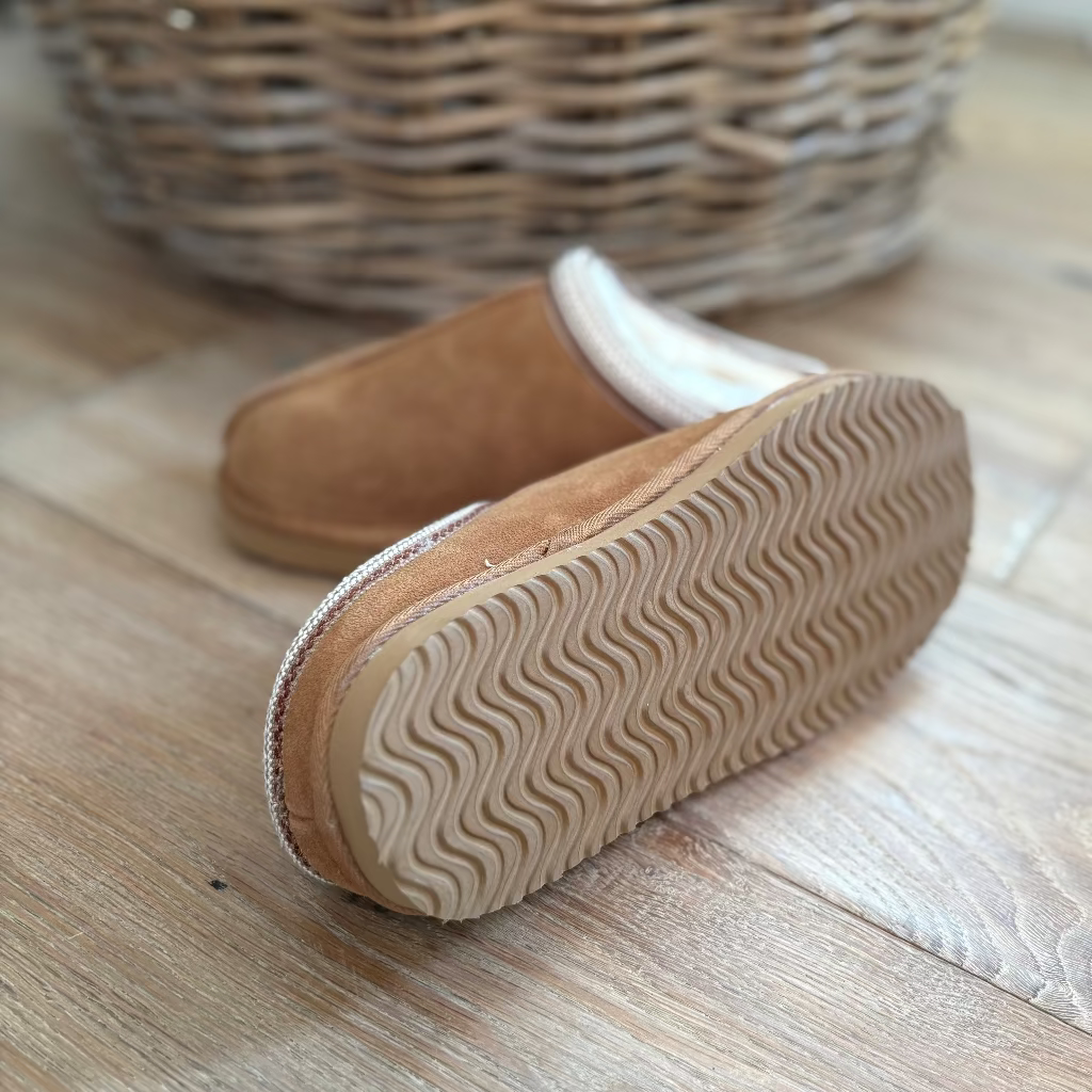 A pair of tan suede slip-on slippers with a cream-colored inner lining and a wavy treaded sole, displayed on a light wood floor with a wicker basket in the background.