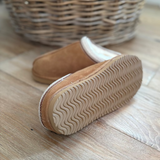 A pair of tan suede slip-on slippers with a cream-colored inner lining and a wavy treaded sole, displayed on a light wood floor with a wicker basket in the background.