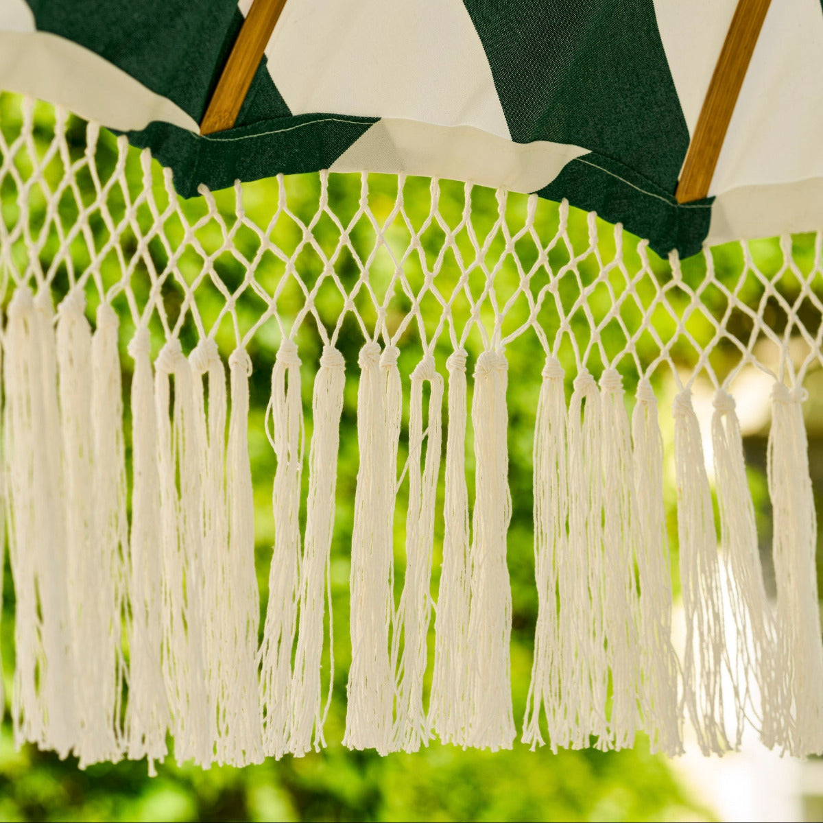 Close-up of a woven patio umbrella canopy with dark green and white diagonal stripes, white fringe tassels, and light wood support poles against a blurred green outdoor background.