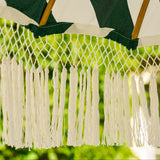 Close-up of a woven patio umbrella canopy with dark green and white diagonal stripes, white fringe tassels, and light wood support poles against a blurred green outdoor background.