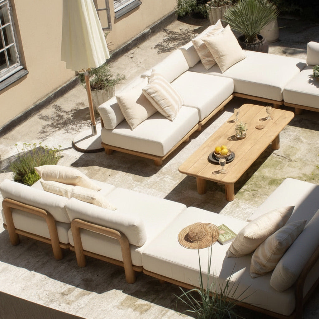 Outdoor patio furniture set with cream cushions and light wood frames, arranged on a stone terrace with a wooden coffee table, potted plants, and a closed umbrella.