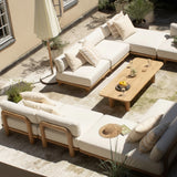 Outdoor patio furniture set with cream cushions and light wood frames, arranged on a stone terrace with a wooden coffee table, potted plants, and a closed umbrella.