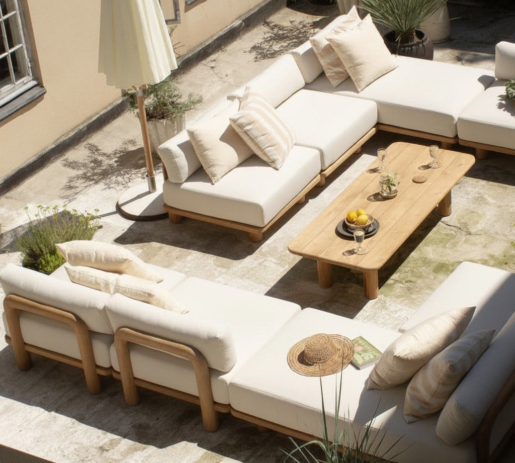 Outdoor patio furniture set with cream cushions and light wood frames, arranged on a stone terrace with a wooden coffee table, potted plants, and a closed umbrella.