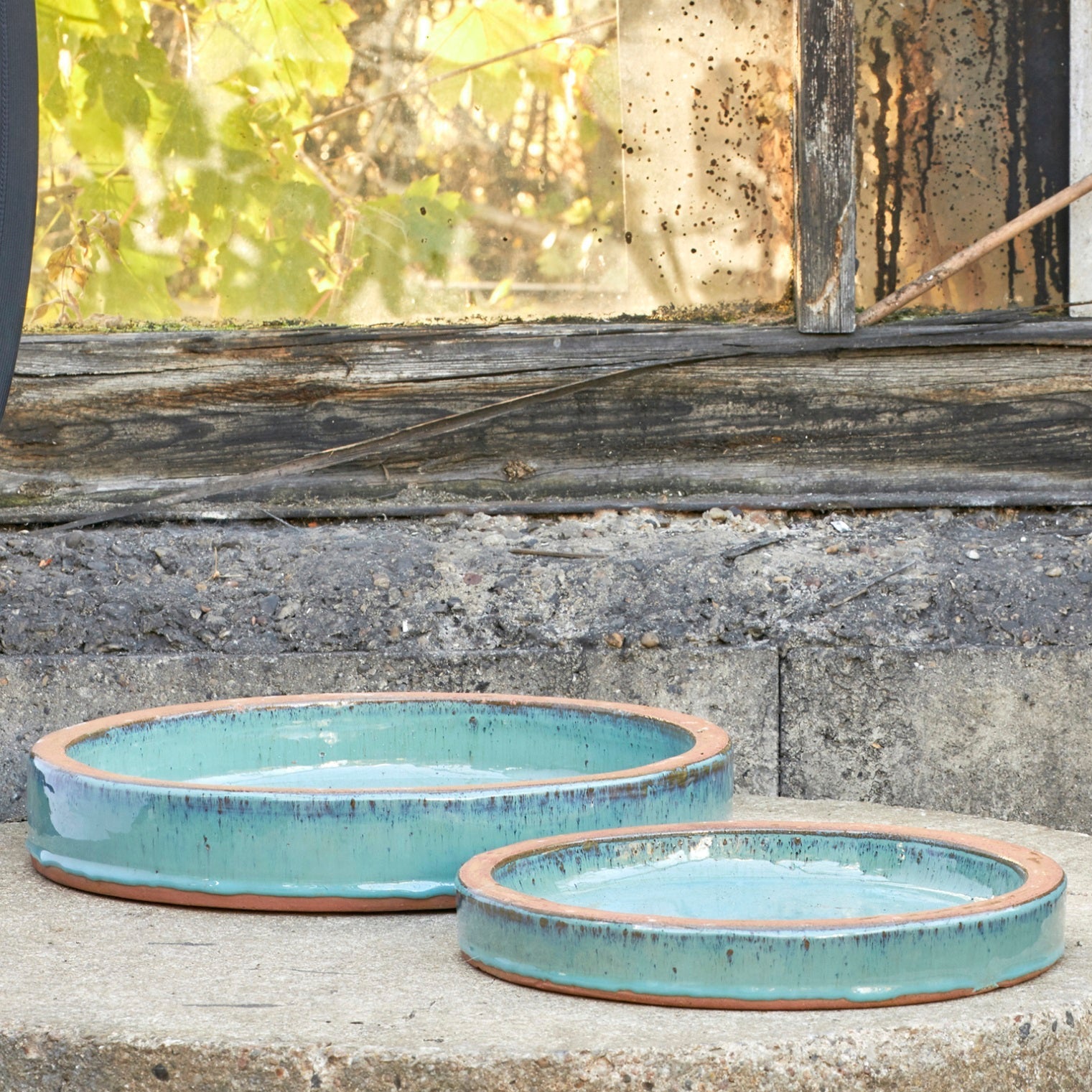 Two round shallow ceramic bird baths with a light blue-green glazed finish and a rustic terracotta rim, displayed on a stone ledge in front of an old weathered window.