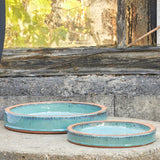 Two round shallow ceramic bird baths with a light blue-green glazed finish and a rustic terracotta rim, displayed on a stone ledge in front of an old weathered window.