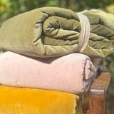 Three rolled velvet bedding pieces stacked on a wooden chair outdoors: a mustard yellow top roll, a pale pink middle roll, and an olive green bottom roll, with a blurred green garden background.