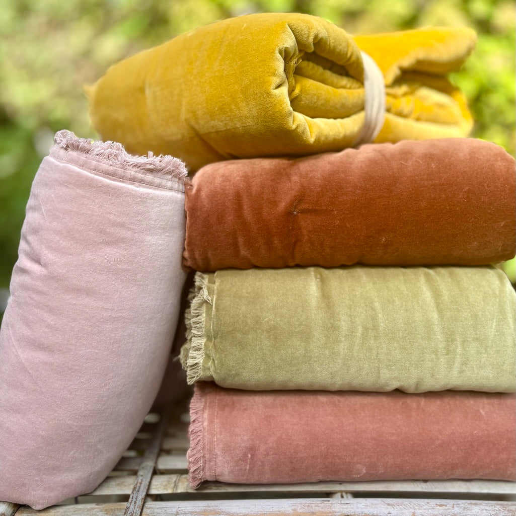 Four folded velvet fabric pieces in dusty pink, rust orange, sage green, and mustard yellow, with one pink piece standing upright beside the stack on a rustic wooden surface against a blurred green outdoor background.
