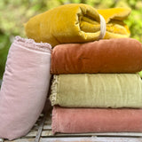 Four folded velvet fabric pieces in dusty pink, rust orange, sage green, and mustard yellow, with one pink piece standing upright beside the stack on a rustic wooden surface against a blurred green outdoor background.