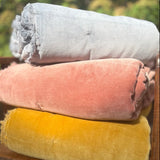 Three rolled velvet throw blankets in light gray, dusty pink, and mustard yellow stacked on a wooden chair with a blurred outdoor garden background.
