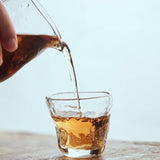 Glass teapot pouring tea into a glass cup on a wooden surface with a light blue background
