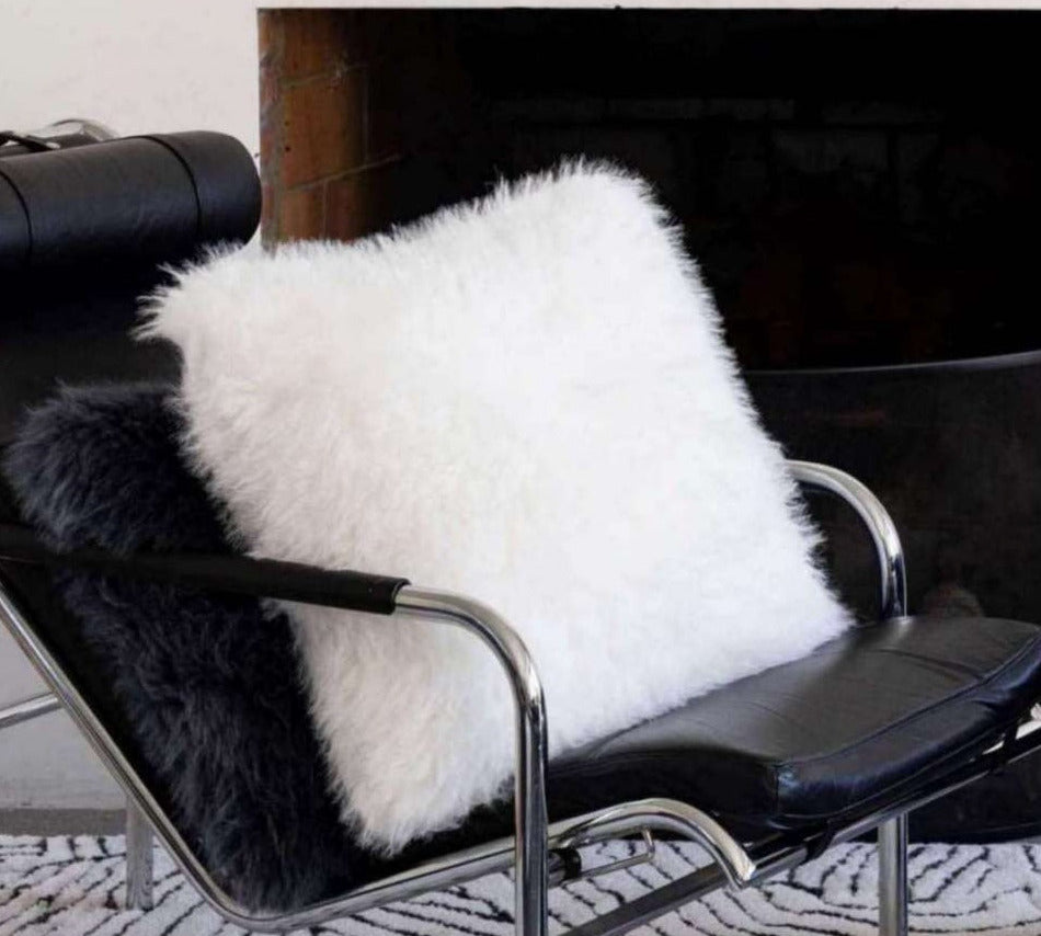 A fluffy white faux fur throw cushion and a black faux fur throw pillow resting on a black leather lounge chair in front of a fireplace.