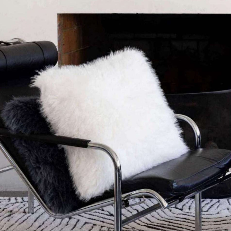 A fluffy white faux fur throw cushion and a black faux fur throw pillow resting on a black leather lounge chair in front of a fireplace.