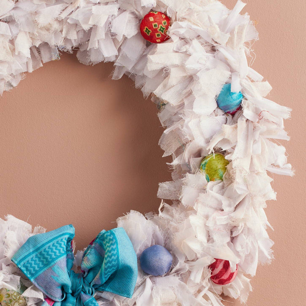 A handmade white fabric wreath decorated with colorful patterned ornaments and a large blue bow, hanging against a solid peach-colored wall.