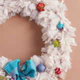 A handmade white fabric wreath decorated with colorful patterned ornaments and a large blue bow, hanging against a solid peach-colored wall.