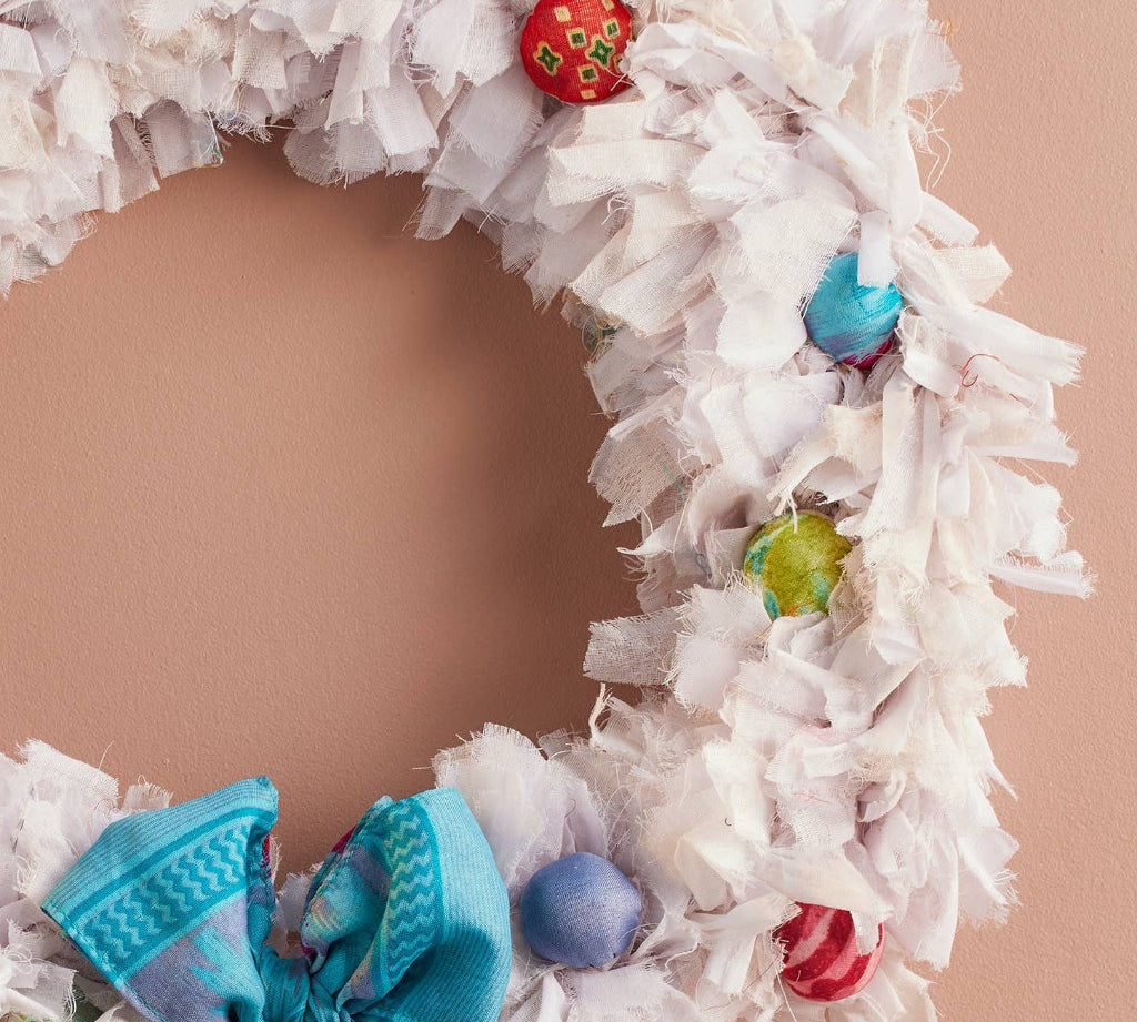 A handmade white fabric wreath decorated with colorful patterned ornaments and a large blue bow, hanging against a solid peach-colored wall.