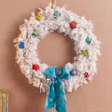 A white fringed Christmas wreath hanging on a dusty pink wall above a wooden mantel. The wreath is decorated with colorful glass ornaments and a bright turquoise blue knitted bow at the bottom center.