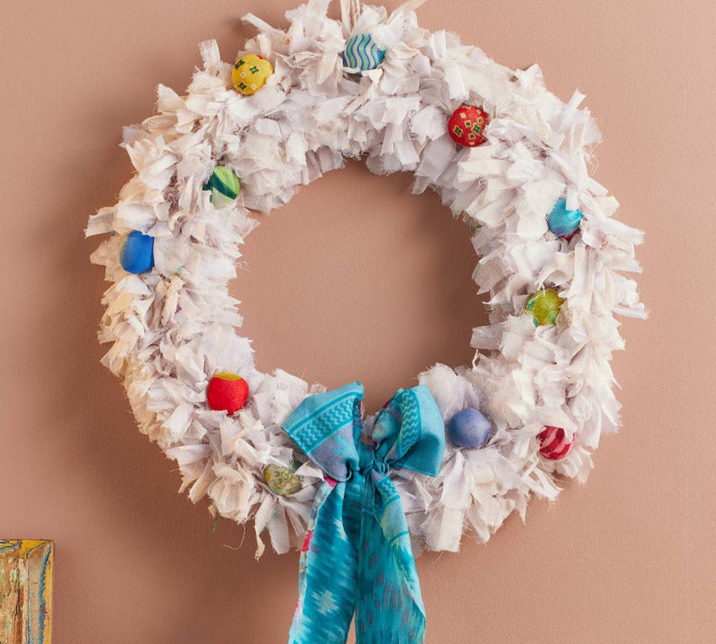 A white fringed Christmas wreath hanging on a dusty pink wall above a wooden mantel. The wreath is decorated with colorful glass ornaments and a bright turquoise blue knitted bow at the bottom center.